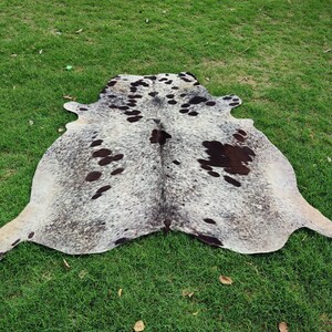 May include: A natural cowhide rug featuring a speckled gray and white base with irregular dark brown and black patches distributed across the surface. The hide has an organic, asymmetrical shape with distinct edges and a textured hair-on-hide finish.