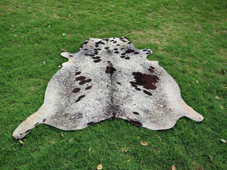 May include: A natural cowhide rug featuring a speckled gray and white base with irregular dark brown and black patches distributed across the surface. The hide has an organic, asymmetrical shape with distinct edges and a textured hair-on-hide finish.