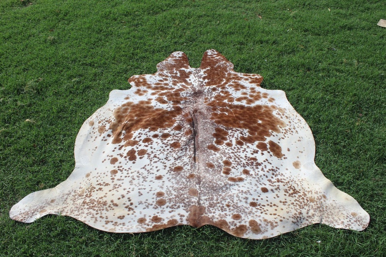 May include: A white cowhide rug with brown spots. The rug is laid out on a green grassy surface.