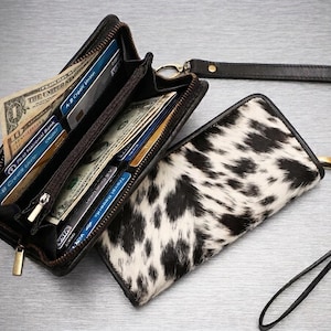 May include: A black leather wallet with a cow print hair-on-hide exterior, featuring a zip closure, a wrist strap, and multiple interior slots containing several credit cards and United States paper currency notes.