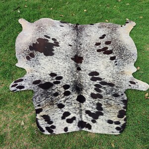 May include: A natural cowhide rug featuring a speckled pattern of black and dark brown spots against a white and grey background. The hide has an irregular, organic shape with distinct edges and a dense, short-haired texture throughout the surface.