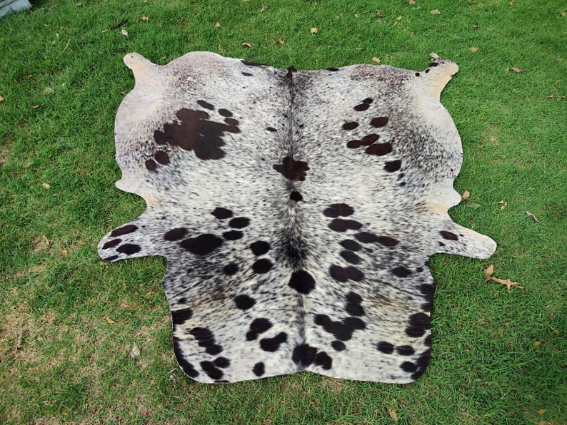 May include: A natural cowhide rug featuring a speckled pattern of black and dark brown spots against a white and grey background. The hide has an irregular, organic shape with distinct edges and a dense, short-haired texture throughout the surface.