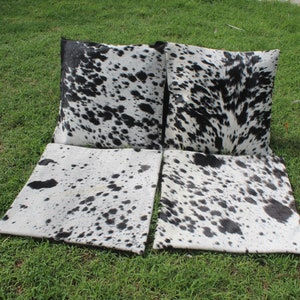 May include: Four black and white cowhide throw pillows with a black and white cowhide pattern. The pillows are arranged in a row on a green grassy surface.