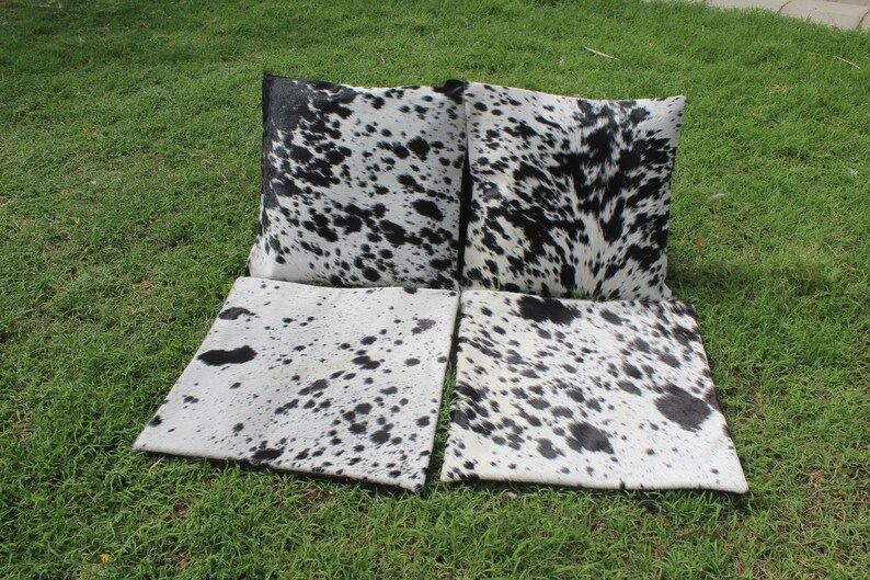 May include: Four black and white cowhide throw pillows with a black and white cowhide pattern. The pillows are arranged in a row on a green grassy surface.