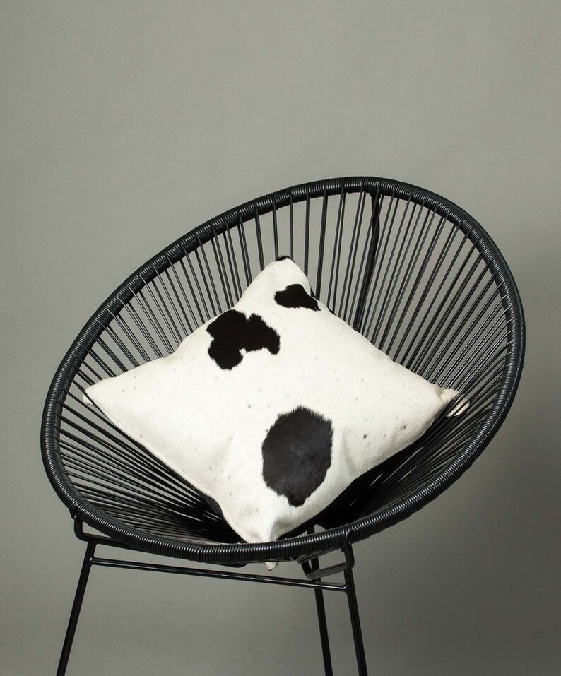 May include: A black and white cowhide pillow on a black wire chair. The pillow has a white background with black spots.