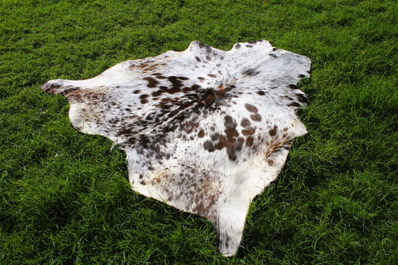 May include: A natural cowhide rug featuring an irregular, organic shape with a white base color and scattered dark brown and black spots. The hide displays a smooth, short-haired texture across its entire surface, showcasing a unique pattern.