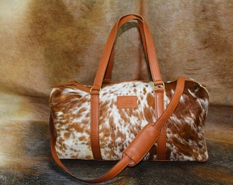Könnte beinhalten: Eine braune Ledertasche mit einem weißen und braunen Kuhfellmuster. Die Tasche hat einen langen Schultergurt und zwei Tragegriffe oben.