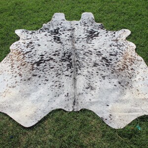 May include: A natural cowhide rug featuring a salt and pepper pattern with a mix of white, grey, black, and light brown spots. The hide has an irregular, organic shape with a central seam and a textured surface, displaying varied color density.