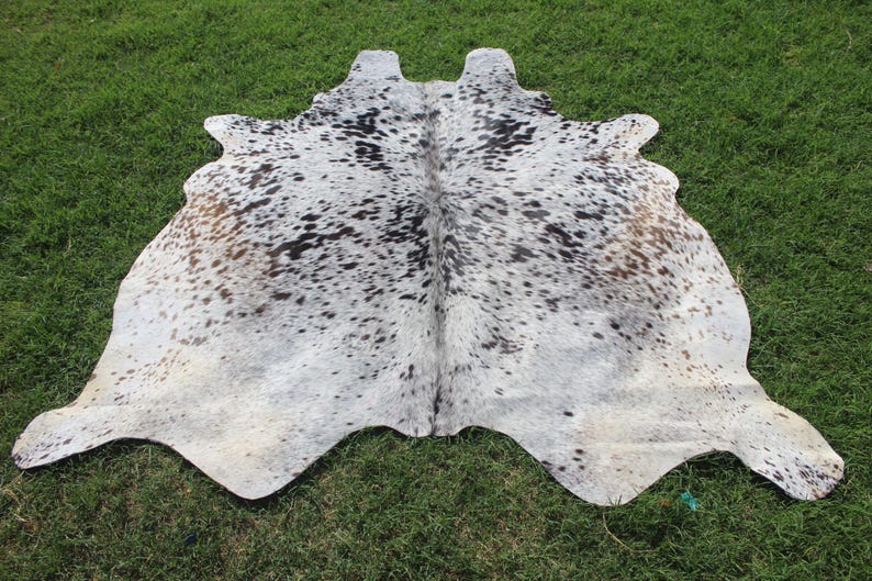 May include: A natural cowhide rug featuring a salt and pepper pattern with a mix of white, grey, black, and light brown spots. The hide has an irregular, organic shape with a central seam and a textured surface, displaying varied color density.