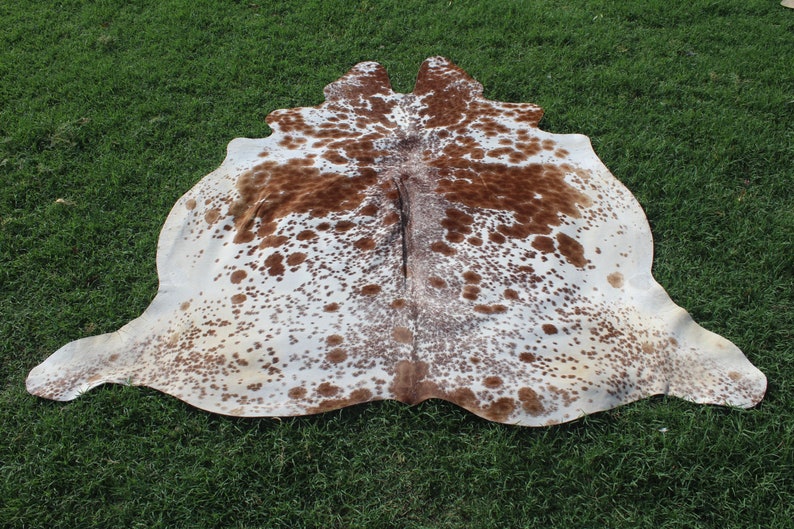 May include: A brown and white cowhide rug with a spotted pattern. The rug is laid out on a green grassy surface.