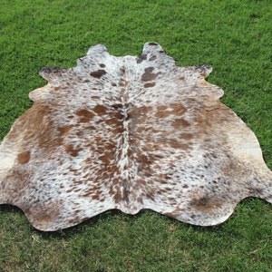 May include: A brown and white cowhide rug with a spotted pattern. The rug is laid out on a green grassy surface.