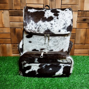 以下が含まれることがあります： 黒と白の牛革のバックパックで、ジッパー付きのコンパートメントが2つあります。バックパックは柔らかくしなやかな革で作られており、ユニークな自然なパターンを持っています。