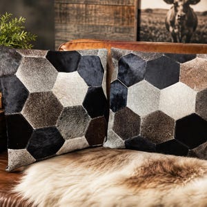 Peut inclure: Deux coussins carrés présentant un motif géométrique composé de pièces hexagonales. Les pièces sont en peau avec poils dans des tons variés de noir, brun foncé, brun clair, gris et blanc, créant un aspect texturé et multicolore.