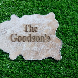 May include: A white cowhide rug with the text "The Goodson's" engraved on it. The rug is on a green artificial grass background.