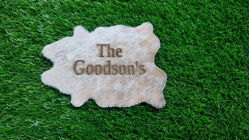 May include: A white cowhide rug with the text "The Goodson's" engraved on it. The rug is on a green artificial grass background.