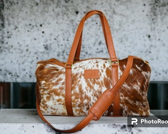 Könnte beinhalten: Eine braune Leder-Reisetasche mit einem weißen und braunen Kuhfellmuster. Die Tasche hat zwei obere Griffe und einen langen, verstellbaren Schultergurt.