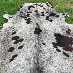 May include: A natural cowhide rug featuring a speckled pattern of black and dark brown spots against a white and grey background. The hide has an irregular, organic shape with a central seam and distinct patches of dark fur across the surface.