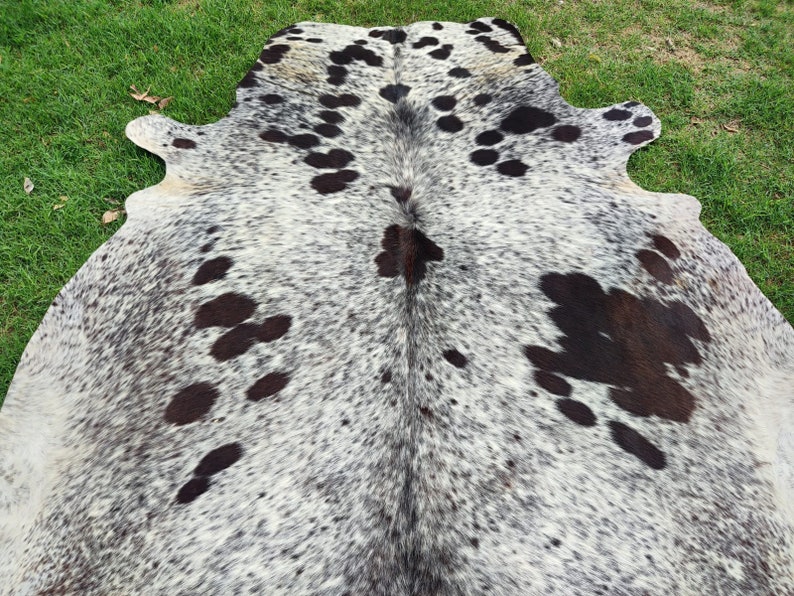 May include: A natural cowhide rug featuring a speckled pattern of black and dark brown spots against a white and grey background. The hide has an irregular, organic shape with a central seam and distinct patches of dark fur across the surface.