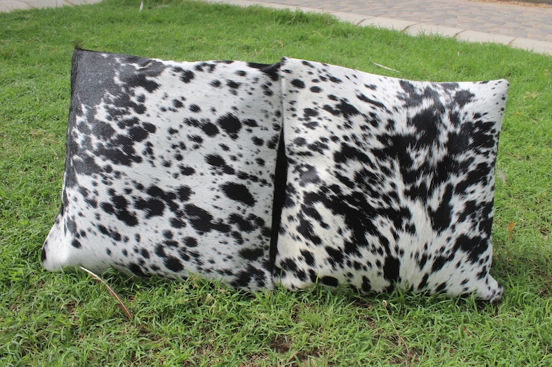 May include: Two black and white cowhide throw pillows with a spotted pattern. The pillows are on a green grassy surface.