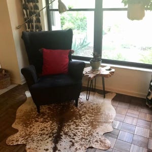 Może przedstawiać: Czarny fotel z czerwoną poduszką stoi na brązowym i białym dywanie ze sk&oacute;ry krowiej przed oknem. Mały drewniany stolik pomocniczy z czarnymi n&oacute;żkami w kształcie szpilek stoi obok fotela.
