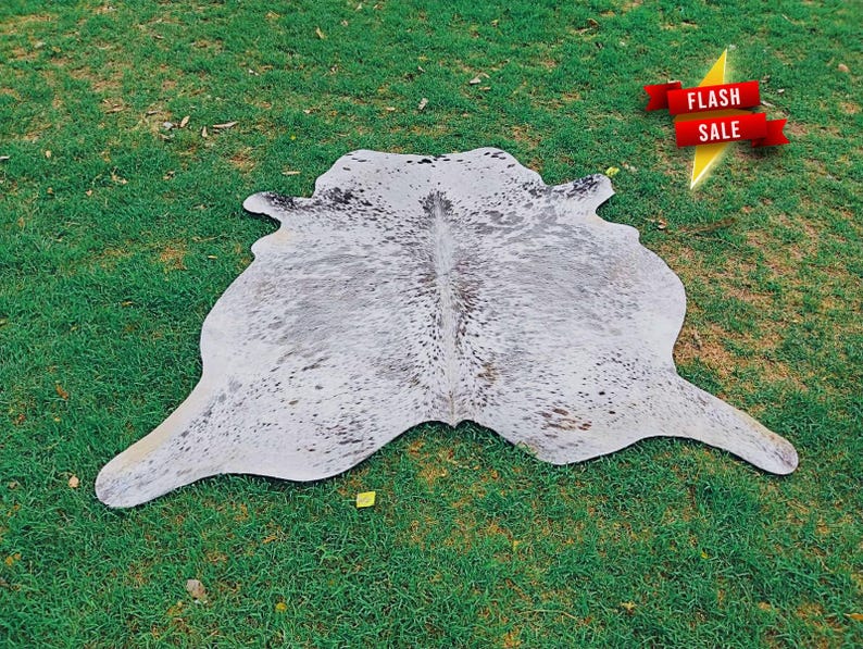 May include: A cowhide rug featuring a natural, irregular shape with a white base color and scattered grey and black speckles throughout the surface. A red banner with the white text FLASH SALE is positioned in the upper right corner of the image.