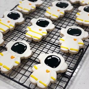 Puede incluir: Una rejilla de enfriamiento con nueve galletas de azúcar decoradas con forma de astronautas. Las galletas son blancas con caras negras, rayas amarillas y corazones naranjas.