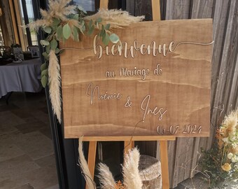 May include: A wooden sign with the text "Bienvenue au Mariage de Noémie & Ines 06-07-2024" in white lettering. The sign is decorated with dried pampas grass and greenery.