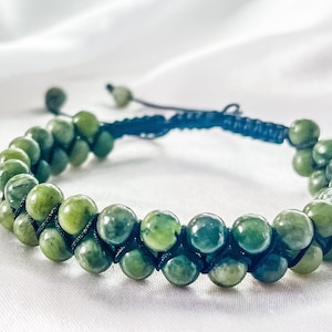 May include: A green stone beaded bracelet with a black cord. The bracelet is made of round beads and has a knotted closure.