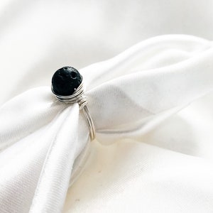 May include: A handcrafted ring featuring a dark, textured lava rock bead, securely wrapped with silver-colored wire. The ring's band is a simple, polished silver. The ring is set against a backdrop of white fabric, highlighting its minimalist design.