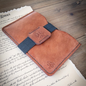 Puede incluir: Una cartera de cuero marrón con cierre de banda elástica negra. La cartera tiene un diseño cosido y un logotipo que dice "PILC".