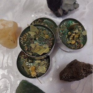 May include: Four small, round, green candles with dried herbs and glitter are arranged with various stones on a white, textured surface. The candles are in silver containers. The stones are in shades of brown, green, and yellow.