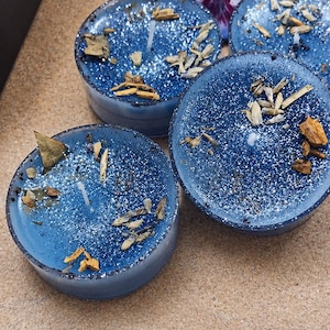 May include: Four blue candles with glitter and dried botanicals. Each candle is round and set in a dark blue container. A purple and white flower is in the background.