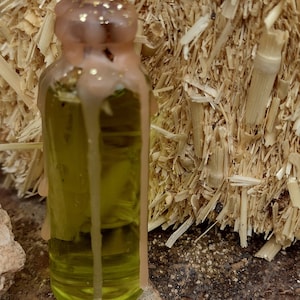 May include: A small glass bottle filled with a yellow liquid, sealed with a pinkish-tan wax cap adorned with gold flecks. The bottle is set against a backdrop of straw and a textured surface.