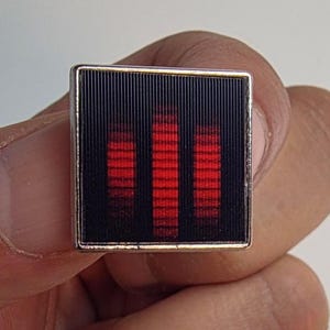 Könnte beinhalten: Quadratischer Manschettenknopf mit schwarzem Hintergrund und drei vertikalen roten Balken. Der Manschettenknopf hat einen silberfarbenen Rand und wird in einer Hand gehalten. Das Design ähnelt einem Equalizer oder einer Schallwelle.