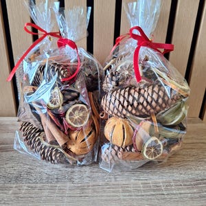 May include: Two clear plastic bags filled with dried potpourri, including brown pinecones, sliced dried citrus fruits, and cinnamon sticks. Each bag is tied at the top with a thin red ribbon, securing the contents inside the transparent packaging.