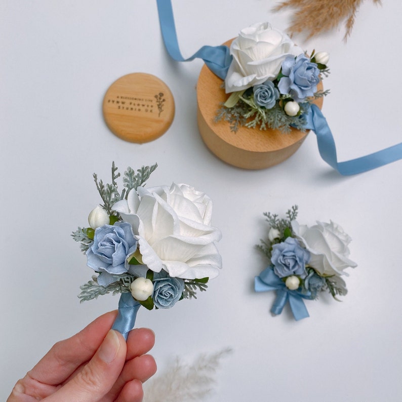 Puede incluir: Tres boutonni&egrave;res con rosas blancas y flores azules. Las boutonni&egrave;res est&aacute;n atadas con una cinta azul. Las boutonni&egrave;res est&aacute;n sobre una superficie blanca.