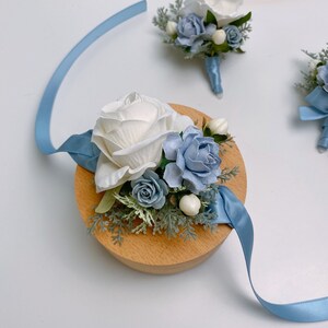 Puede incluir: Un boutonniere de flores blancas y azules con una rosa blanca, flores azules y vegetaci&oacute;n. El boutonniere est&aacute; unido a una cinta azul claro y est&aacute; sentado sobre una base de madera.