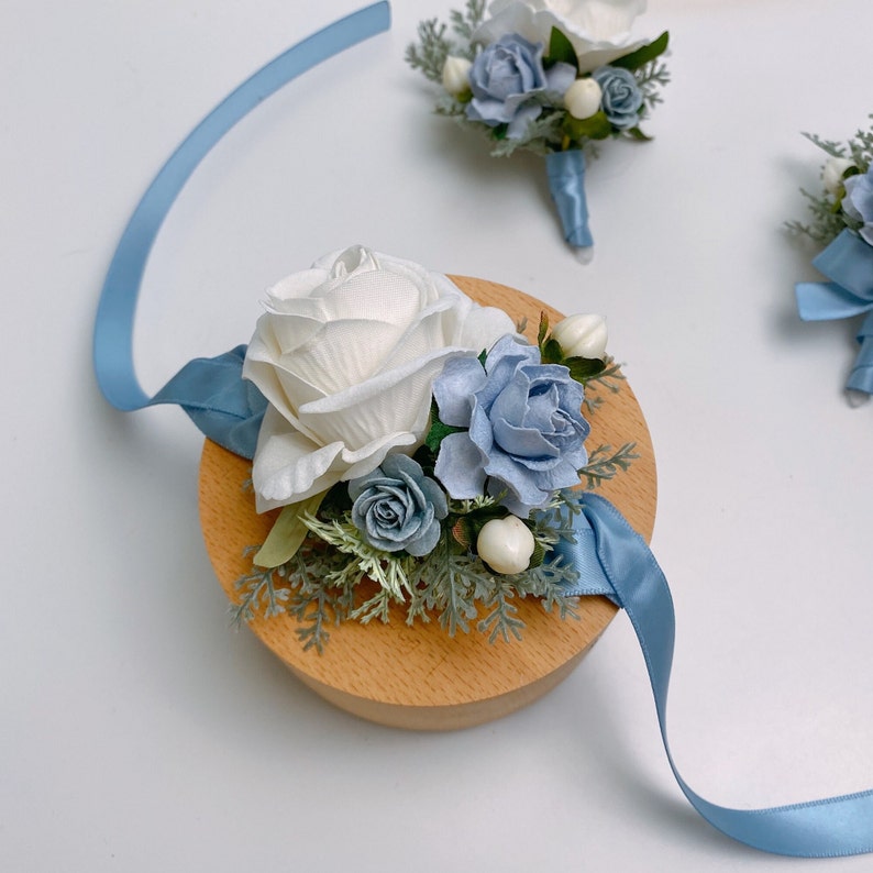 Puede incluir: Un boutonniere de flores blancas y azules con una rosa blanca, flores azules y vegetaci&oacute;n. El boutonniere est&aacute; unido a una cinta azul claro y est&aacute; sentado sobre una base de madera.