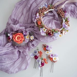 Könnte beinhalten: Eine Sammlung von floralen Haaraccessoires, darunter ein Blumenkranz, ein Kamm und Haarnadeln. Die Stücke zeigen orangefarbene Rosen, weiße Gänseblümchen und lila und rosa Blüten. Sie sind auf einem lavendelfarbenen Stoff angeordnet.