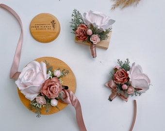 Puede incluir: Tres boutonnières florales de color rosa y rubor con vegetación sobre bases de madera. Las boutonnières están dispuestas sobre una superficie blanca con una cinta rosa.
