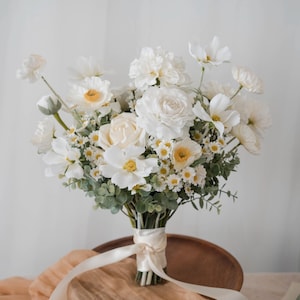 May include: A white bridal bouquet featuring white roses, daisies, and other white blooms. The bouquet is tied with a white satin ribbon.