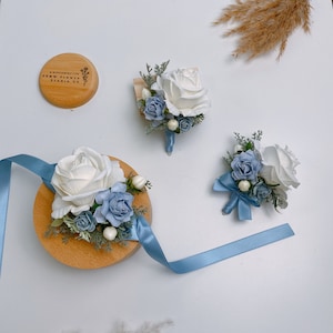 May include: Three white and blue flower buttonholes with a blue ribbon. The buttonholes are on a wooden surface. The text "A Blooming Life, FYNH FLOWER STUDIO UK" is on a wooden circle.