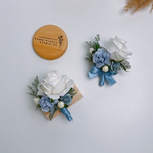 Puede incluir: Dos boutonni&egrave;res de flores blancas y azules con una base de madera y una cinta azul. Las boutonni&egrave;res est&aacute;n sobre una superficie blanca. Un c&iacute;rculo de madera con el texto "A Blossoming Life FYNW FLOWER STUDIO UK" tambi&eacute;n est&aacute; sobre la superficie.