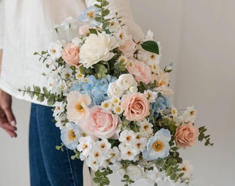 Könnte beinhalten: Ein Blumenstrauß aus künstlichen Blumen in den Farben Hellrosa, Creme, Weiß und Hellblau. Das Arrangement besteht aus Rosen, Hortensien und kleinen gänseblümchenartigen Blüten, die mit grünen Eukalyptus-Blattstielen durchsetzt sind.