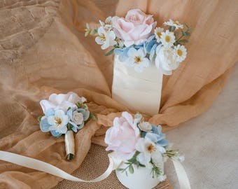 Könnte beinhalten: Eine Auswahl an Blumenarrangements mit pastellrosa Rosen, hellblauen Blüten und weißen Gänseblümchen. Die Arrangements umfassen einen Anstecker, ein Handgelenk-Corsage und ein größeres Mittelstück, alle mit cremefarbenen Bändern.