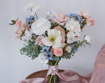 Könnte beinhalten: Ein Blumenstrauß mit weißen, hellrosa und staubblauen Blüten, gemischt mit grünem Eukalyptuslaub. Die Stiele sind zusammengebunden und mit einem hellrosa Satinband versehen, was ein abgerundetes Arrangement verschiedener Blüten ergibt.