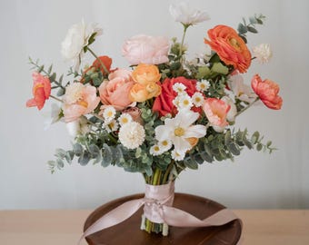 May include: A floral bouquet featuring a mix of orange, peach, pink, and white flowers, including ranunculus, cosmos, and small daisies, accented with green eucalyptus leaves. The stems are bound together with a long, light pink satin ribbon.