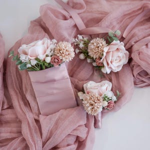Puede incluir: Arreglos florales en color rubor, con rosas y otras flores, dispuestos sobre una tela rosa empolvado. Los arreglos incluyen un ramillete de muñeca, un prendido y un ramillete de bolsillo, perfectos para bodas u ocasiones especiales.