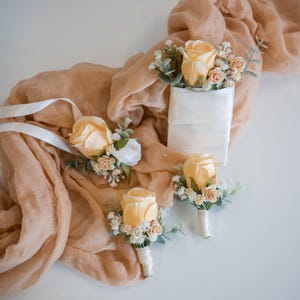 May include: A collection of floral accessories featuring peach-coloured roses, small white filler flowers, and green eucalyptus leaves. The set includes a wrist corsage with a white ribbon and three boutonnieres wrapped in white satin ribbon.