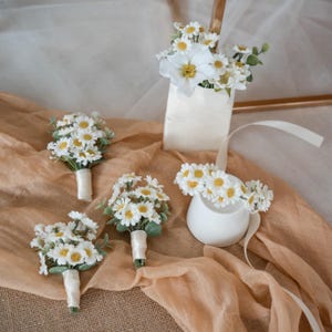 May include: A selection of white daisy floral arrangements. The image shows several small bouquets, a floral crown, and a larger arrangement in a white container. The daisies have yellow centres and green leaves, and the bouquets are wrapped with white ribbon.